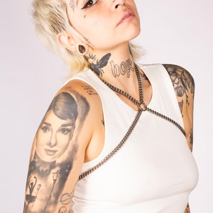 May include: A woman with short blonde hair and numerous tattoos, wearing a white vest top with a silver chain harness. The word "hope" is tattooed on her neck.