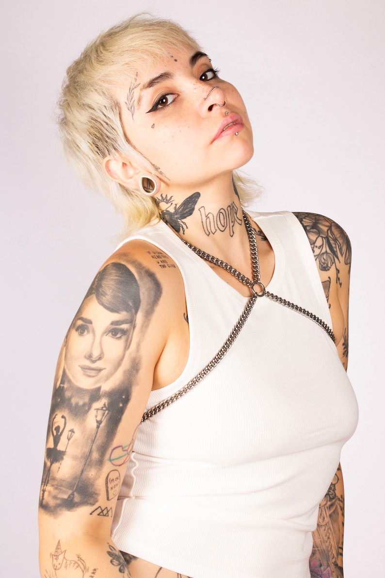 May include: A woman with short blonde hair and numerous tattoos, wearing a white vest top with a silver chain harness. The word "hope" is tattooed on her neck.