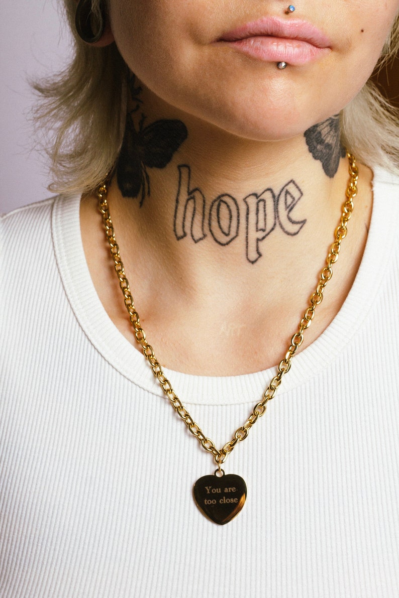 Op de afbeelding: Goudkleurige ketting met een hartvormige hanger. De hanger heeft de gegraveerde tekst "You are too close". Het woord "hope" is op de nek getatoe&euml;erd.