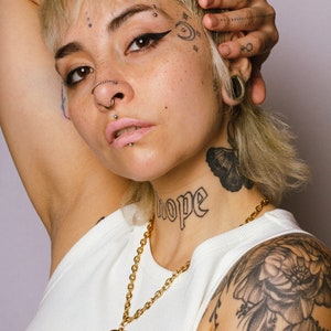 Op de afbeelding: Een persoon draagt een witte tanktop en een gouden ketting met een hartvormige hanger. Het woord "hope" is op de nek getatoe&euml;erd. De persoon heeft meerdere gezichtspiercings en tatoeages op het gezicht en de arm.