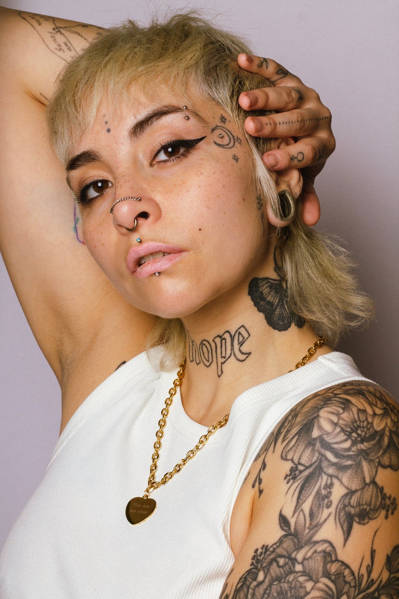 Op de afbeelding: Een persoon draagt een witte tanktop en een gouden ketting met een hartvormige hanger. Het woord "hope" is op de nek getatoe&euml;erd. De persoon heeft meerdere gezichtspiercings en tatoeages op het gezicht en de arm.