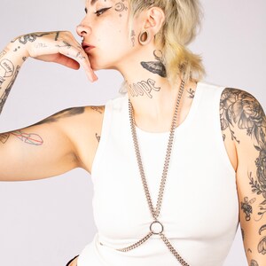Puede incluir: Una persona con el pelo corto rubio y tatuajes lleva un top blanco sin mangas con un arn&eacute;s de cuerpo de cadena plateada. La cadena tiene un anillo grande en el centro.