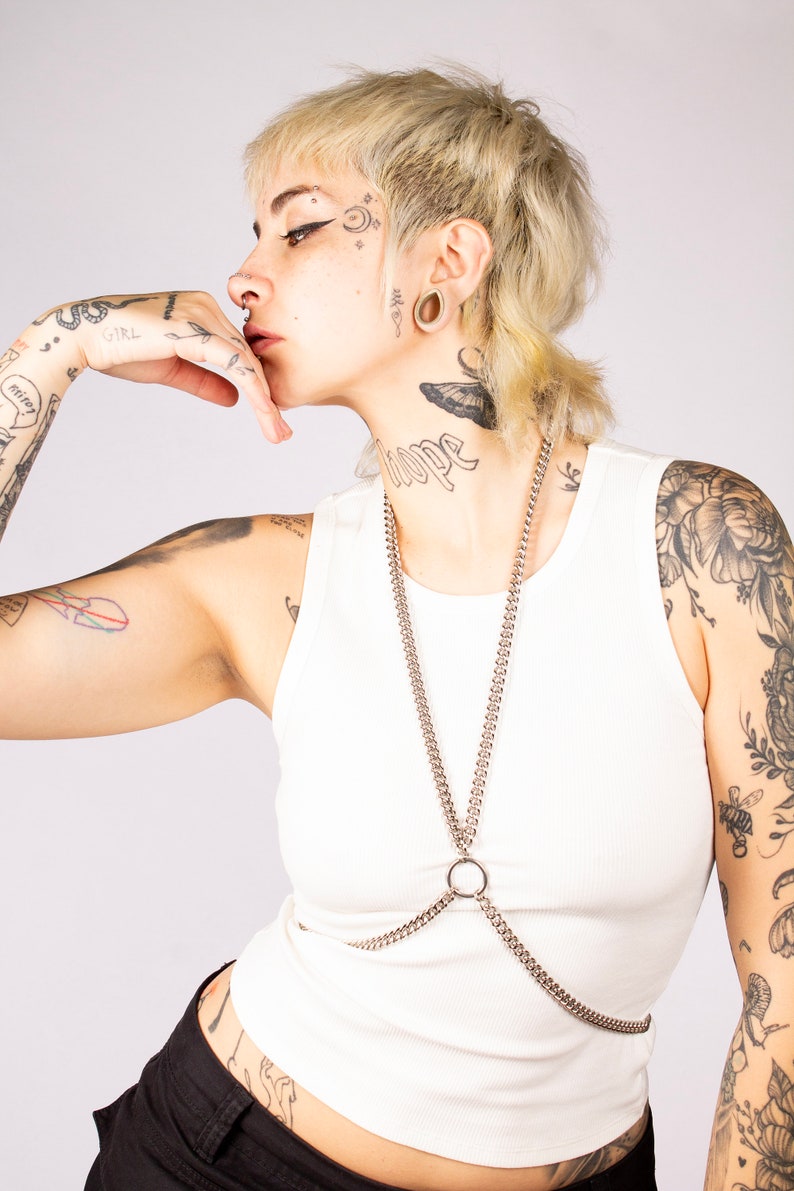 Puede incluir: Una persona con el pelo corto rubio y tatuajes lleva un top blanco sin mangas con un arn&eacute;s de cuerpo de cadena plateada. La cadena tiene un anillo grande en el centro.