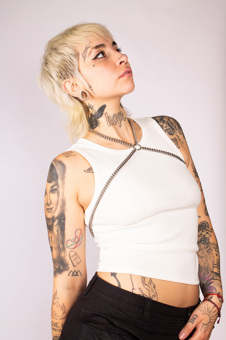 May include: A person with short blonde hair and numerous tattoos is wearing a white vest top with a silver chain harness and black trousers. The person has the word "hope" tattooed on their neck.