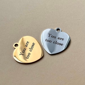 Op de afbeelding: Twee hartvormige hangers, &eacute;&eacute;n goudkleurig en &eacute;&eacute;n zilverkleurig, elk met een klein gaatje aan de bovenkant. De gouden hanger heeft de tekst "You are too close" in zwart gegraveerd. De zilveren hanger heeft dezelfde tekst in zwart gegraveerd.