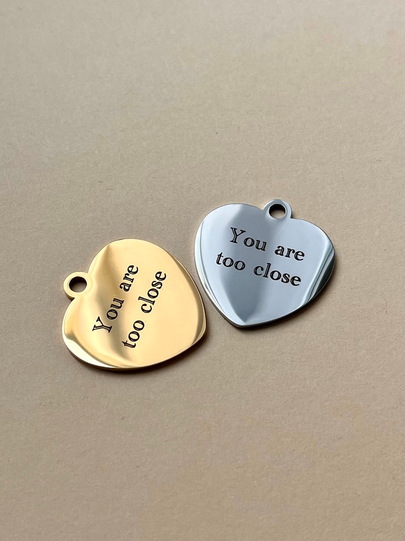 Op de afbeelding: Twee hartvormige hangers, &eacute;&eacute;n goudkleurig en &eacute;&eacute;n zilverkleurig, elk met een klein gaatje aan de bovenkant. De gouden hanger heeft de tekst "You are too close" in zwart gegraveerd. De zilveren hanger heeft dezelfde tekst in zwart gegraveerd.