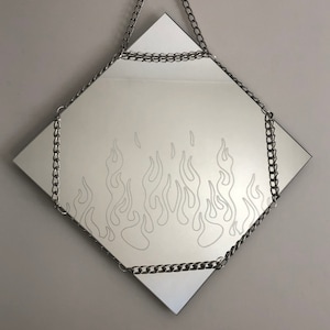 Op de afbeelding: Een ruitvormige spiegel met een zilveren ketting die er bovenaan aan hangt. De spiegel heeft een wit vlammenontwerp dat in het glas is geëtst.