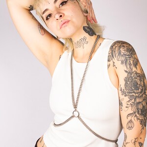 Puede incluir: Una persona con el pelo rubio corto y varios tatuajes lleva un top blanco sin mangas con un arn&eacute;s de cuerpo de cadena plateada y pantalones negros. La persona tiene un tatuaje en el cuello que dice "esperanza".