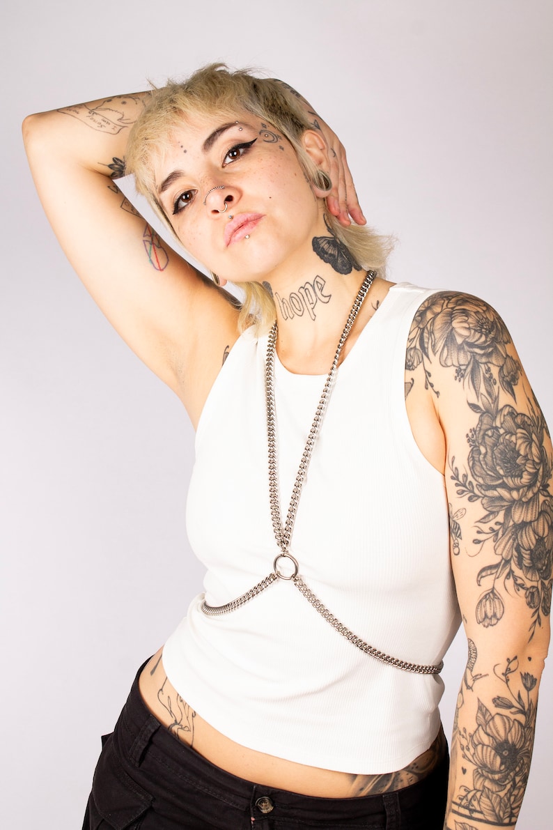 Puede incluir: Una persona con el pelo rubio corto y varios tatuajes lleva un top blanco sin mangas con un arn&eacute;s de cuerpo de cadena plateada y pantalones negros. La persona tiene un tatuaje en el cuello que dice "esperanza".