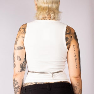 Puede incluir: Un top blanco de tirantes de canal&eacute; con un cintur&oacute;n de cadena plateada. La modelo lleva pantalones negros y tiene tatuajes en los brazos.