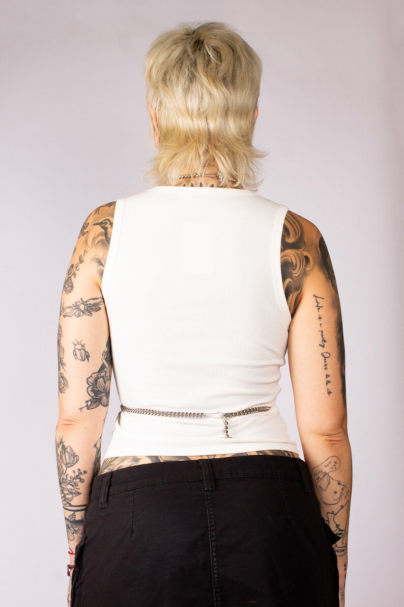 Puede incluir: Un top blanco de tirantes de canal&eacute; con un cintur&oacute;n de cadena plateada. La modelo lleva pantalones negros y tiene tatuajes en los brazos.