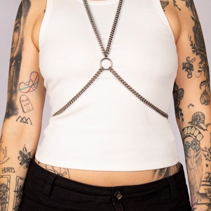 Puede incluir: Un arn&eacute;s de cuerpo de cadena plateada con un anillo grande en el centro. El arn&eacute;s lo lleva una persona con tatuajes en los brazos. La persona lleva una camiseta sin mangas blanca y pantalones negros.