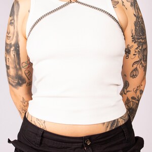 May include: A white tank top with a silver chain body harness. The harness has a large ring in the centre and the word "hope" is tattooed on the model's neck.