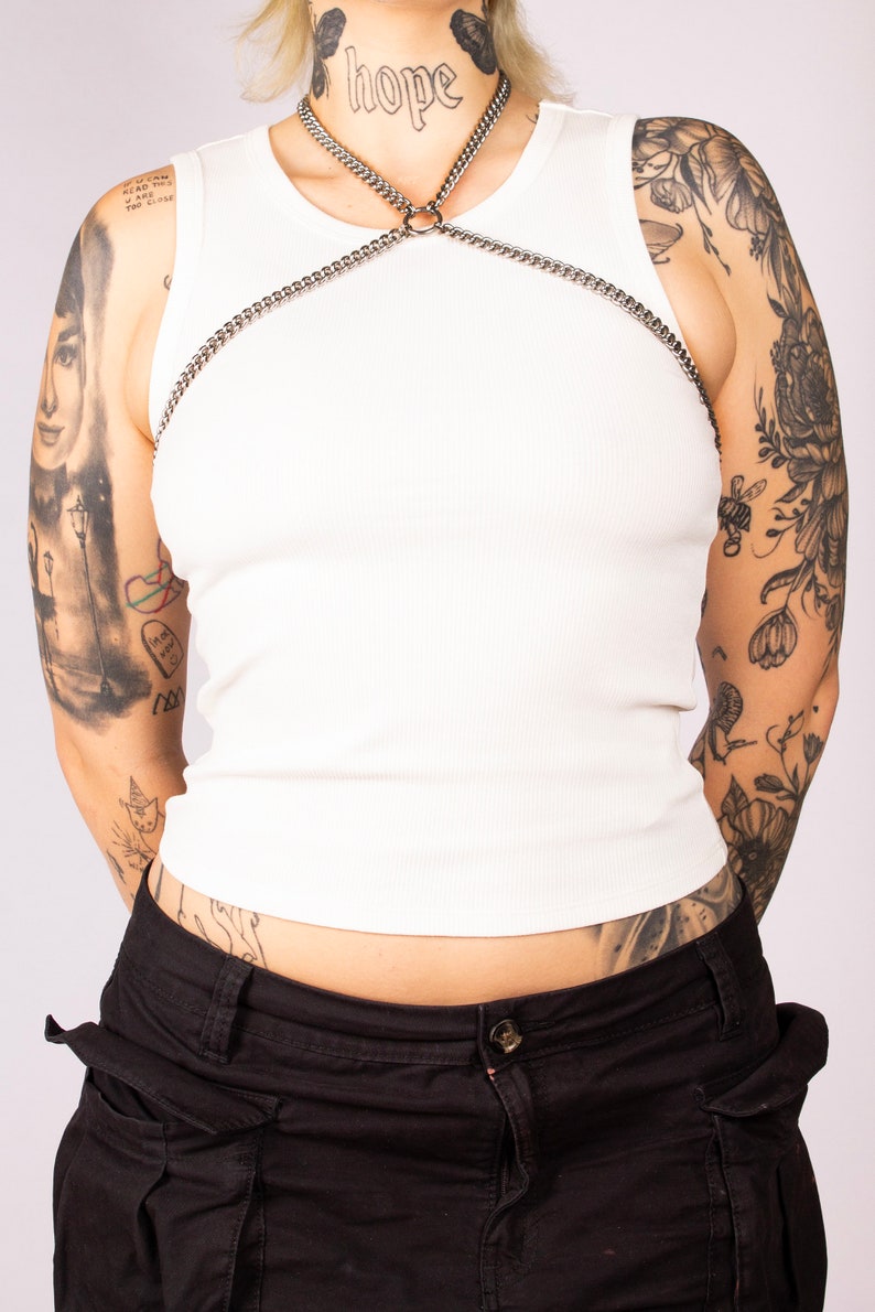 May include: A white tank top with a silver chain body harness. The harness has a large ring in the centre and the word "hope" is tattooed on the model's neck.