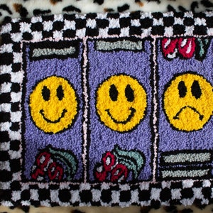Puede incluir: Una alfombra a cuadros blancos y morados con tres caras sonrientes amarillas en el centro. Las caras sonrientes est&aacute;n en fila y la cara sonriente del medio sonr&iacute;e. Las caras sonrientes a cada lado sonr&iacute;en, pero la de la derecha tiene el ce&ntilde;o fruncido. La alfombra tiene un borde rosa y un borde a cuadros blancos y negros.