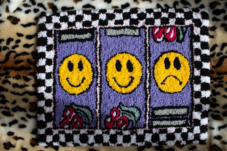 Puede incluir: Una alfombra a cuadros blancos y morados con tres caras sonrientes amarillas en el centro. Las caras sonrientes est&aacute;n en fila y la cara sonriente del medio sonr&iacute;e. Las caras sonrientes a cada lado sonr&iacute;en, pero la de la derecha tiene el ce&ntilde;o fruncido. La alfombra tiene un borde rosa y un borde a cuadros blancos y negros.