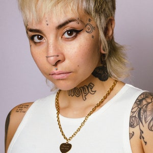 Op de afbeelding: Een gouden ketting met een hartvormige hanger met de tekst "You are too close". De persoon op de foto draagt een witte tanktop en heeft tatoeages op de nek en arm.