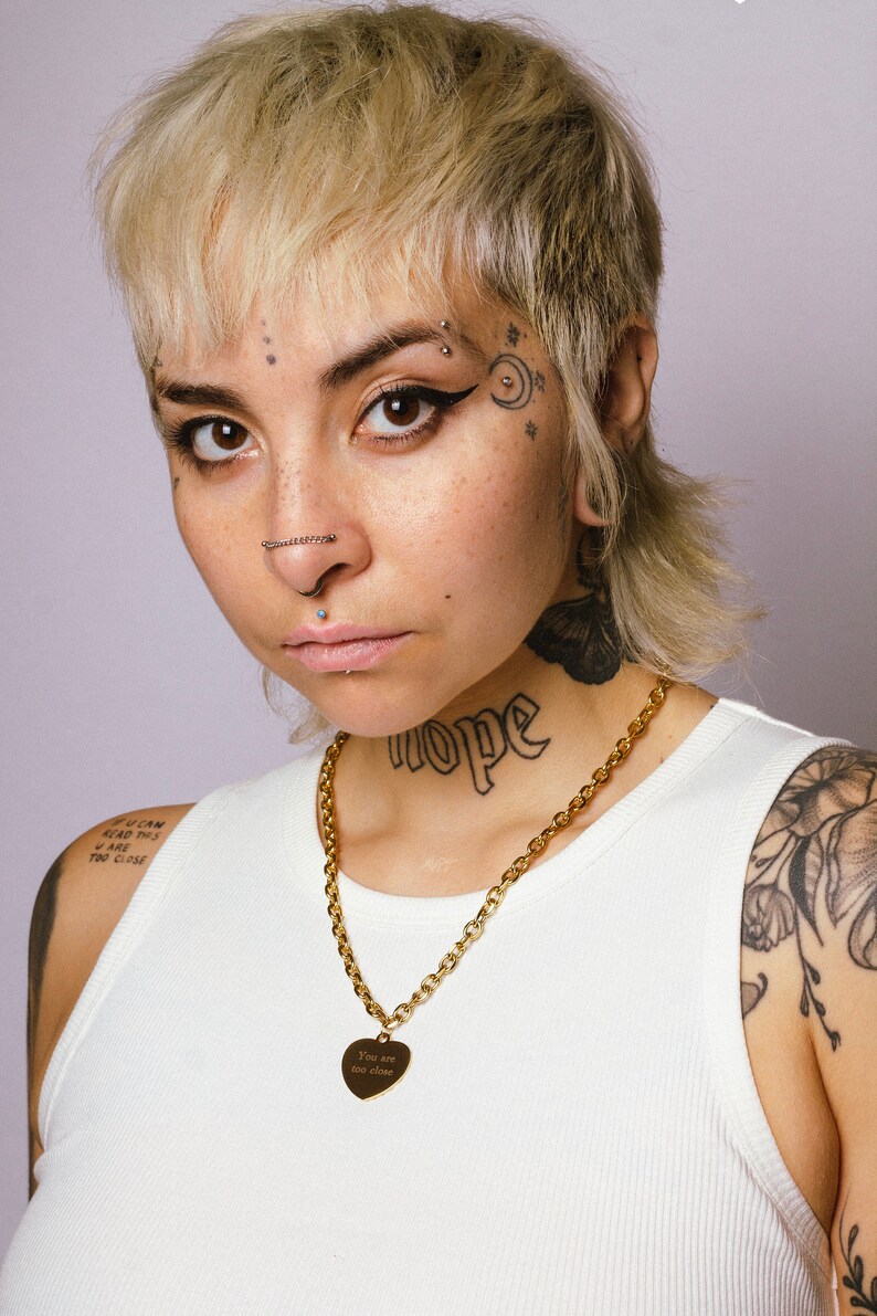 Op de afbeelding: Een gouden ketting met een hartvormige hanger met de tekst "You are too close". De persoon op de foto draagt een witte tanktop en heeft tatoeages op de nek en arm.