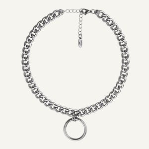 Op de afbeelding: Een zilverkleurige ketting met een grote, cirkelvormige hanger. De ketting heeft een dikke, grove schakelketting en een verstelbare sluiting. De hanger is een eenvoudige, gepolijste ring. De sieraden worden tegen een effen achtergrond getoond.