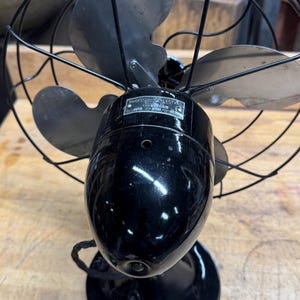 May include: Vintage Emerson Electric fan with a black finish. The fan has three blades and a protective wire cage. The base is round, and the fan's motor housing is oval-shaped. A label on the motor housing reads "Emerson Electric".