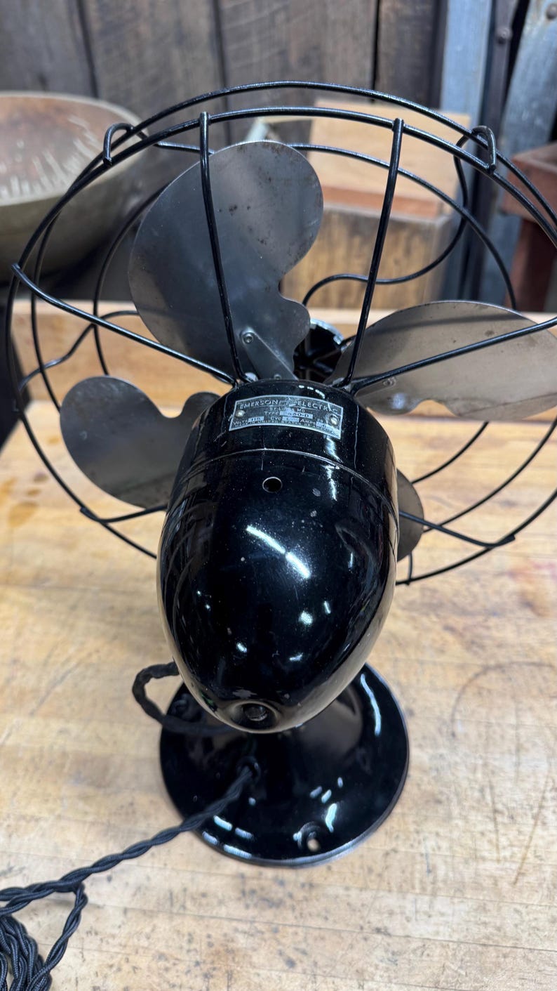 May include: Vintage Emerson Electric fan with a black finish. The fan has three blades and a protective wire cage. The base is round, and the fan's motor housing is oval-shaped. A label on the motor housing reads "Emerson Electric".