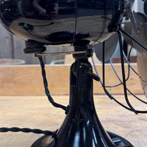 May include: A vintage black electric fan with a shiny finish. The fan has a rounded motor housing, a curved base, and metal blades. The power cord is black and twisted. The fan is likely from the early 20th century.