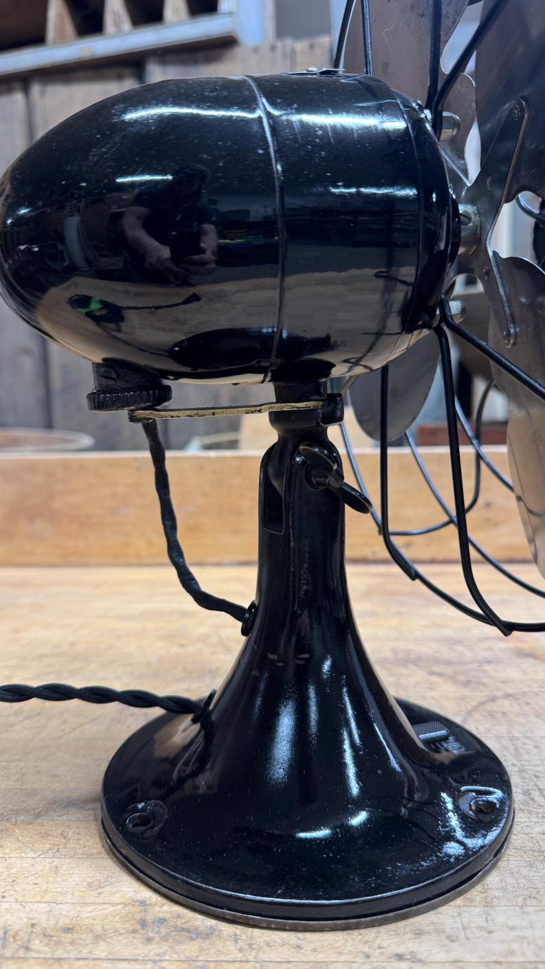 May include: A vintage black electric fan with a shiny finish. The fan has a rounded motor housing, a curved base, and metal blades. The power cord is black and twisted. The fan is likely from the early 20th century.