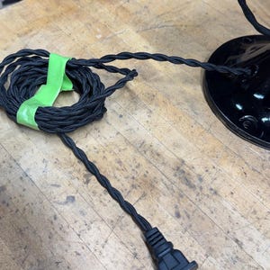 May include: Black electrical cord with a braided fabric covering, coiled and secured with green tape. The cord leads to a black plug and a black lamp base, all on a wooden surface. The cord is approximately 100 inches long.