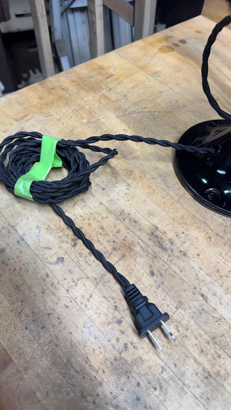 May include: Black electrical cord with a braided fabric covering, coiled and secured with green tape. The cord leads to a black plug and a black lamp base, all on a wooden surface. The cord is approximately 100 inches long.