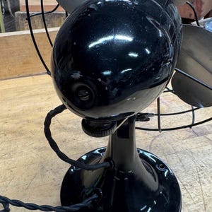 May include: A vintage black electric fan with four blades and a protective cage. The fan has a glossy finish and a round base. The power cord is black and twisted. The fan is sitting on a wooden surface.