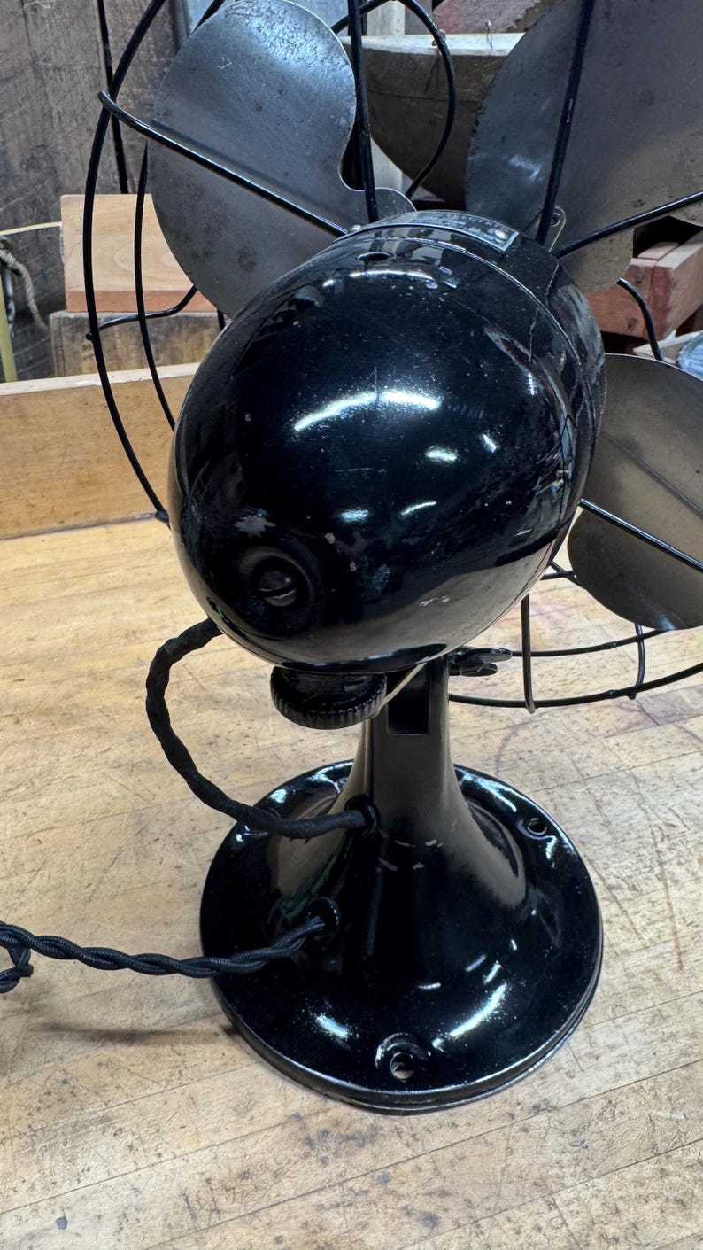 May include: A vintage black electric fan with four blades and a protective cage. The fan has a glossy finish and a round base. The power cord is black and twisted. The fan is sitting on a wooden surface.