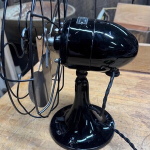 May include: A vintage black electric fan with a rounded motor housing and a wire cage. The fan sits on a round base and has a twisted power cord. The blades are visible behind the cage. The fan is on a wooden surface.