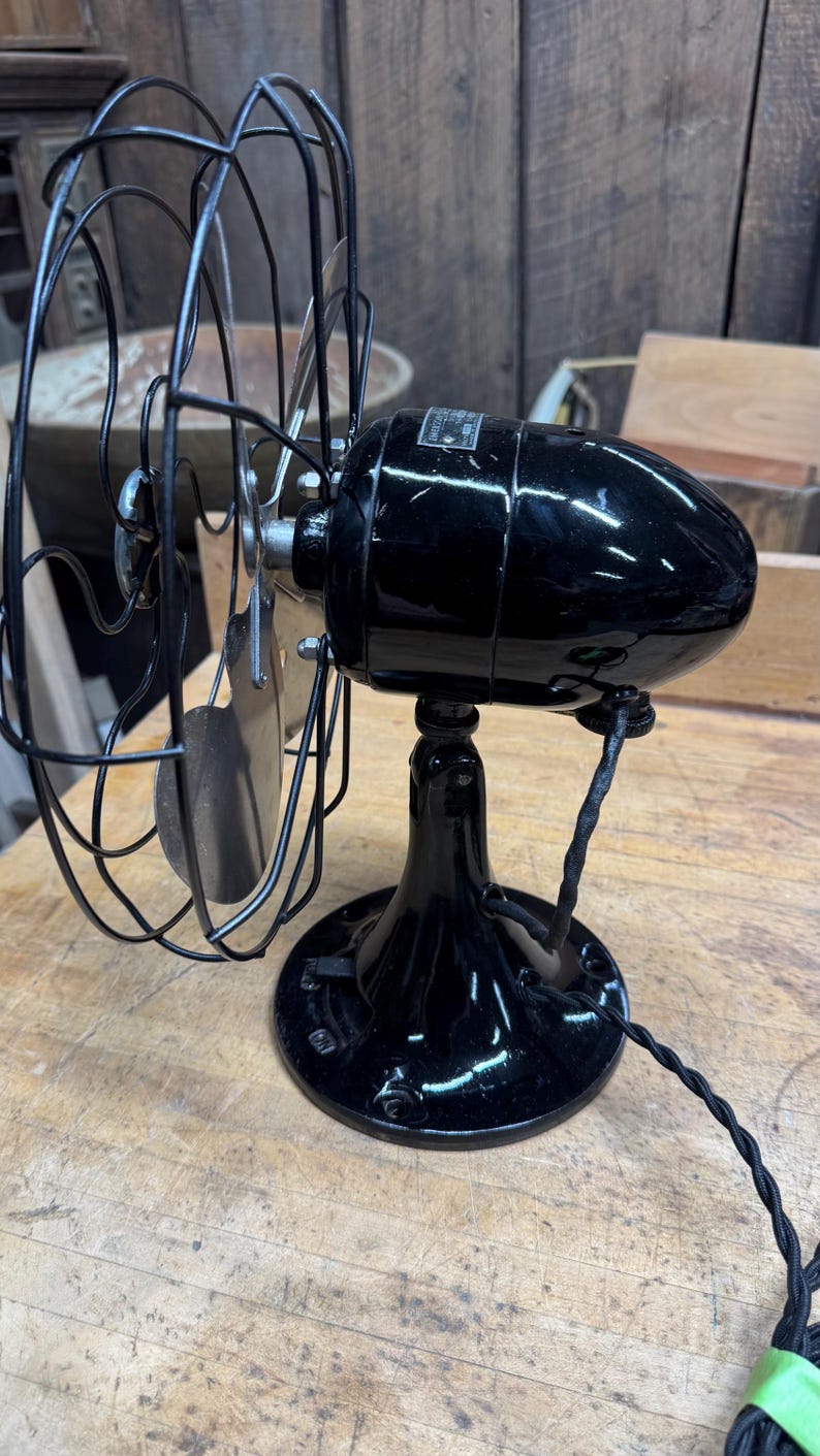 May include: A vintage black electric fan with a rounded motor housing and a wire cage. The fan sits on a round base and has a twisted power cord. The blades are visible behind the cage. The fan is on a wooden surface.