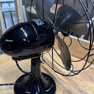 May include: An antique black electric fan with a cage and three blades. The fan has a glossy finish and sits on a round base. The blades are silver and the cage is black wire. The fan is on a wooden surface.