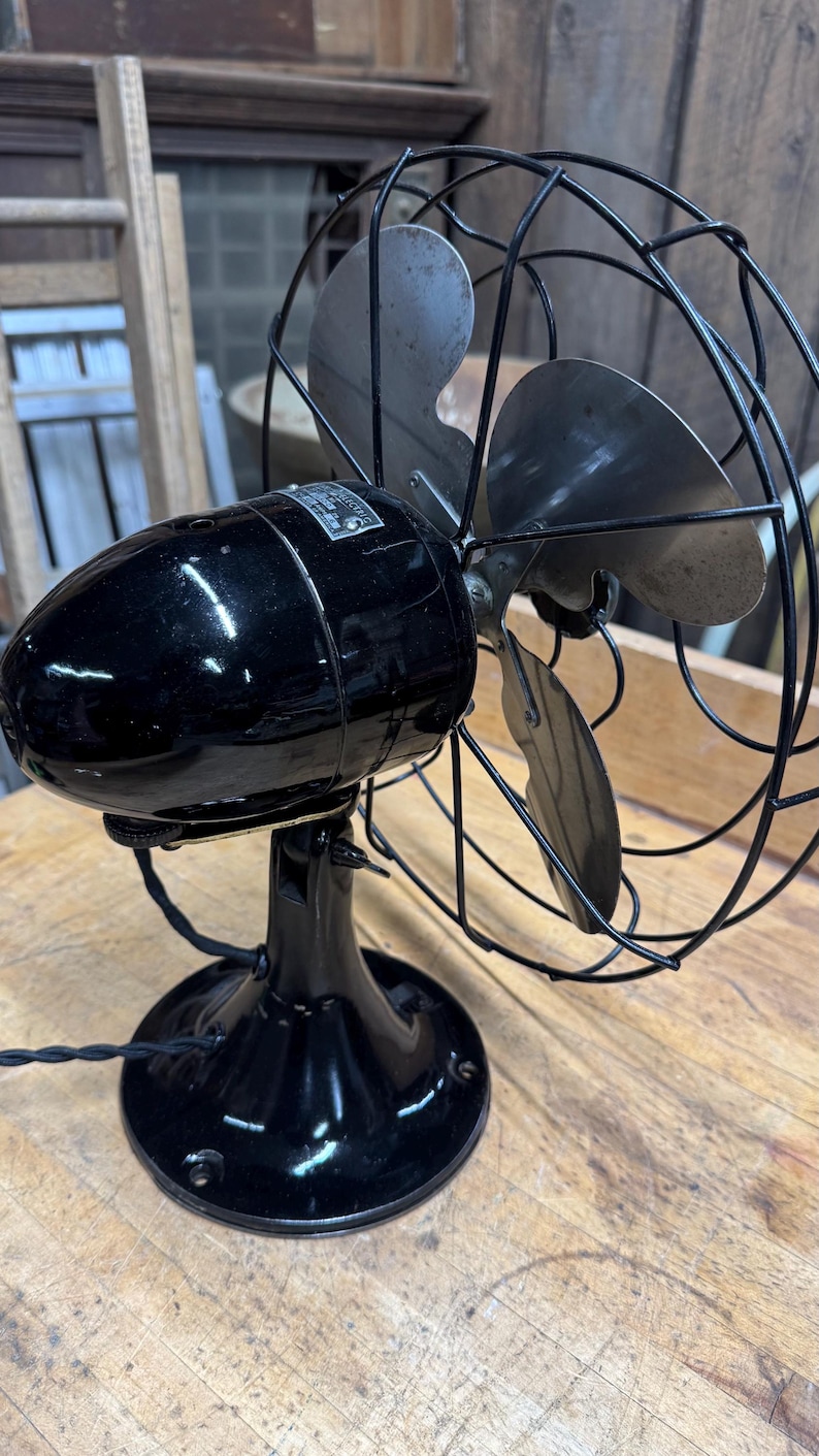 May include: An antique black electric fan with a cage and three blades. The fan has a glossy finish and sits on a round base. The blades are silver and the cage is black wire. The fan is on a wooden surface.