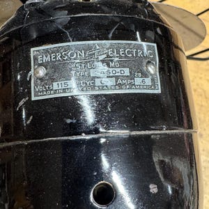 May include: Close-up of a vintage Emerson Electric fan. The black motor housing has a metal plate with text including "EMERSON ELECTRIC", "ST. LOUIS, MO", and "MADE IN UNITED STATES OF AMERICA". The fan blades are silver.