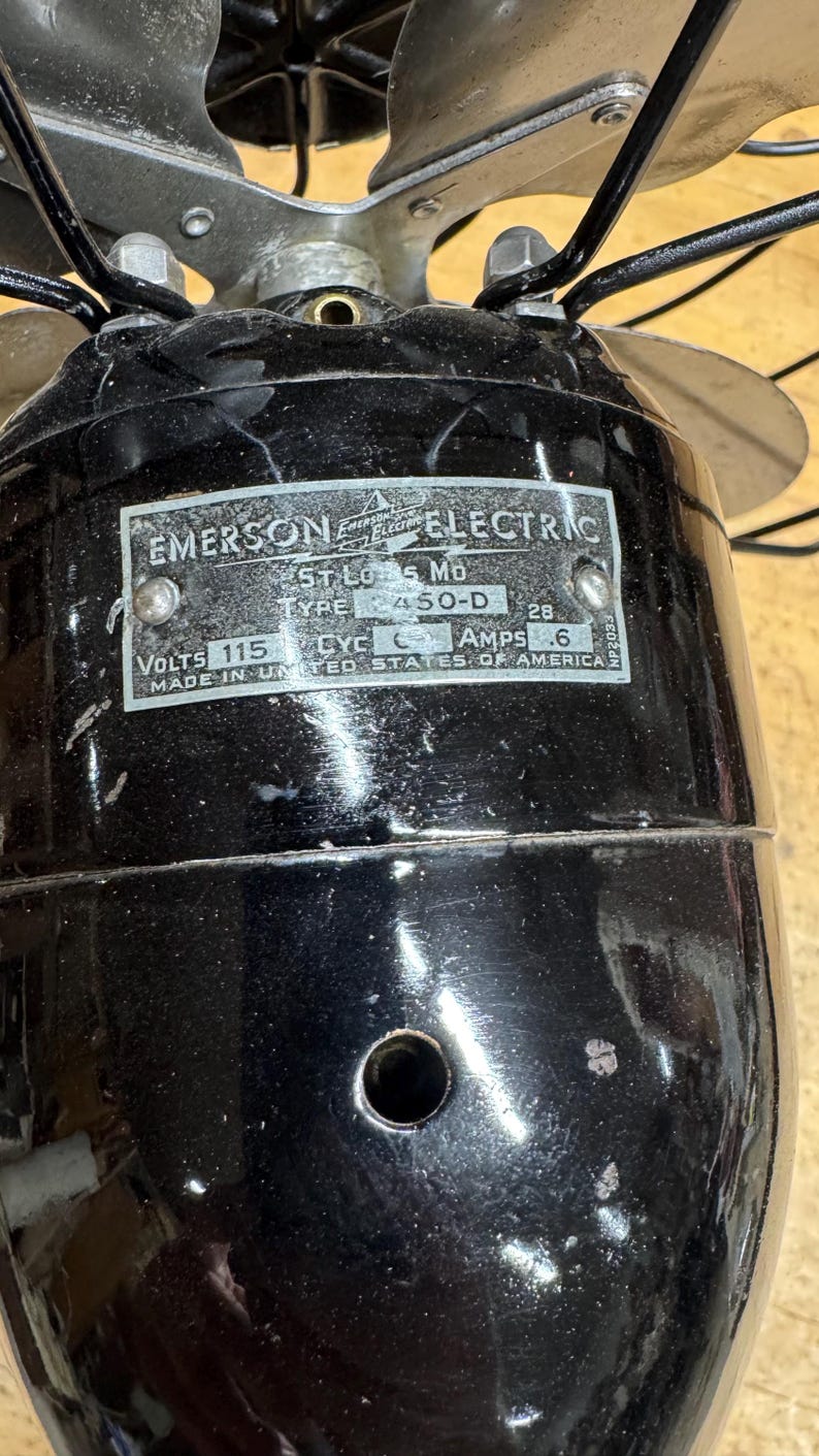 May include: Close-up of a vintage Emerson Electric fan. The black motor housing has a metal plate with text including "EMERSON ELECTRIC", "ST. LOUIS, MO", and "MADE IN UNITED STATES OF AMERICA". The fan blades are silver.