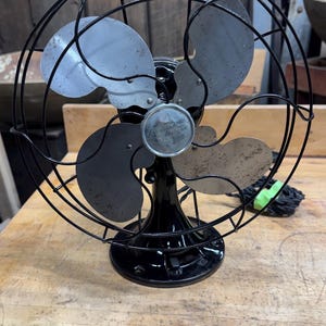 May include: An antique black electric fan with a circular cage and four blades. The fan has a black base and a silver emblem in the center. The blades are a mix of silver and black. The fan is sitting on a wooden surface.