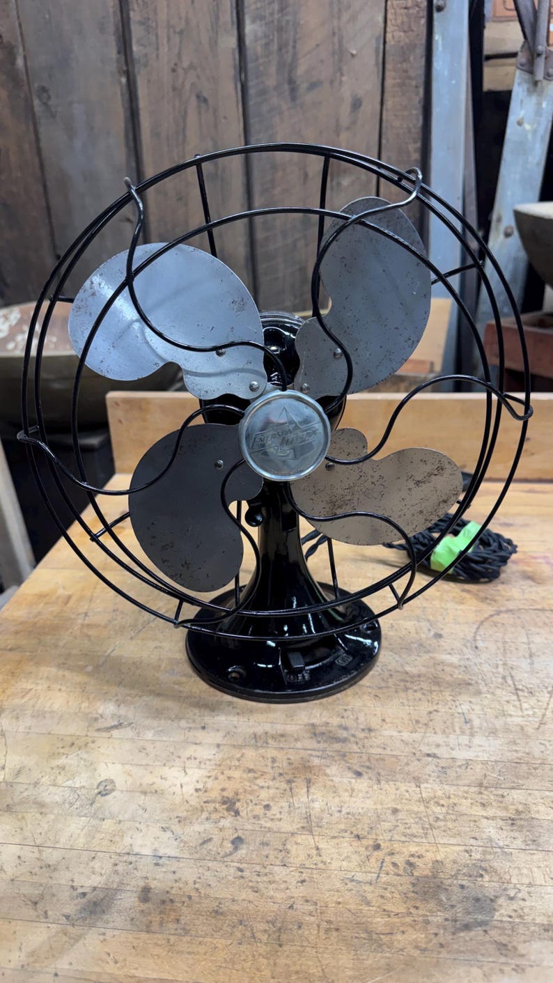 May include: An antique black electric fan with a circular cage and four blades. The fan has a black base and a silver emblem in the center. The blades are a mix of silver and black. The fan is sitting on a wooden surface.