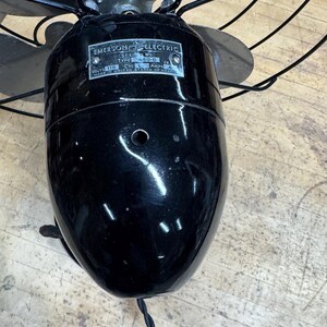 May include: Vintage Emerson Electric oscillating fan with a glossy black finish. The fan has four blades and a metal cage. The motor housing features a label with the Emerson Electric logo and model information. A black power cord extends from the base.
