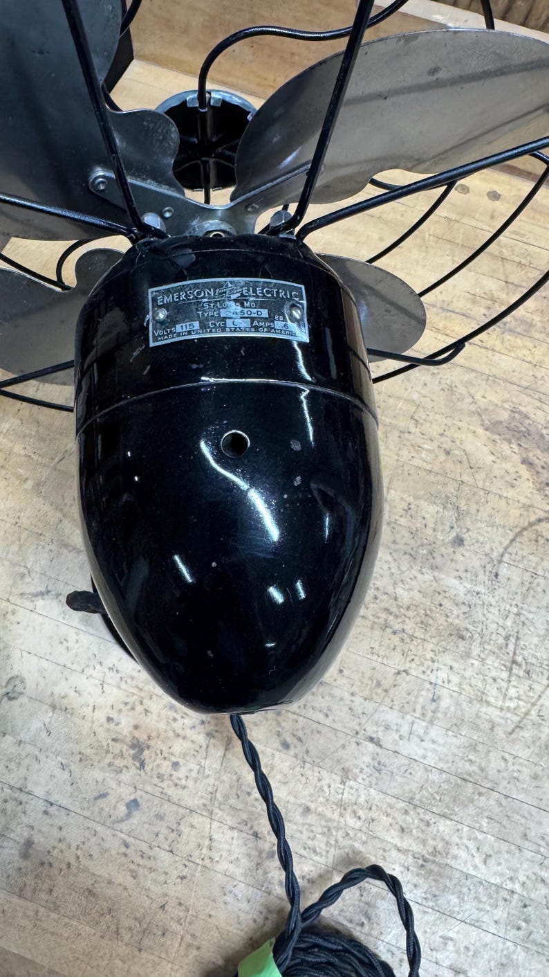 May include: Vintage Emerson Electric oscillating fan with a glossy black finish. The fan has four blades and a metal cage. The motor housing features a label with the Emerson Electric logo and model information. A black power cord extends from the base.