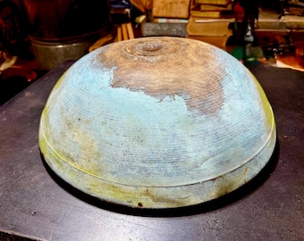May include: A large, antique wooden bowl with a rounded shape. The bowl is painted with a faded blue, brown, and yellow color scheme. The bowl has a circular design and is sitting on a dark surface.