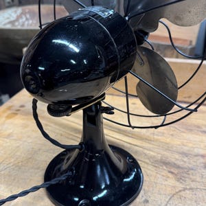 May include: Vintage black electric fan with three silver blades, enclosed by a protective wire cage. The fan has a glossy finish and a round base. A black power cord extends from the base. The fan is a classic home appliance.