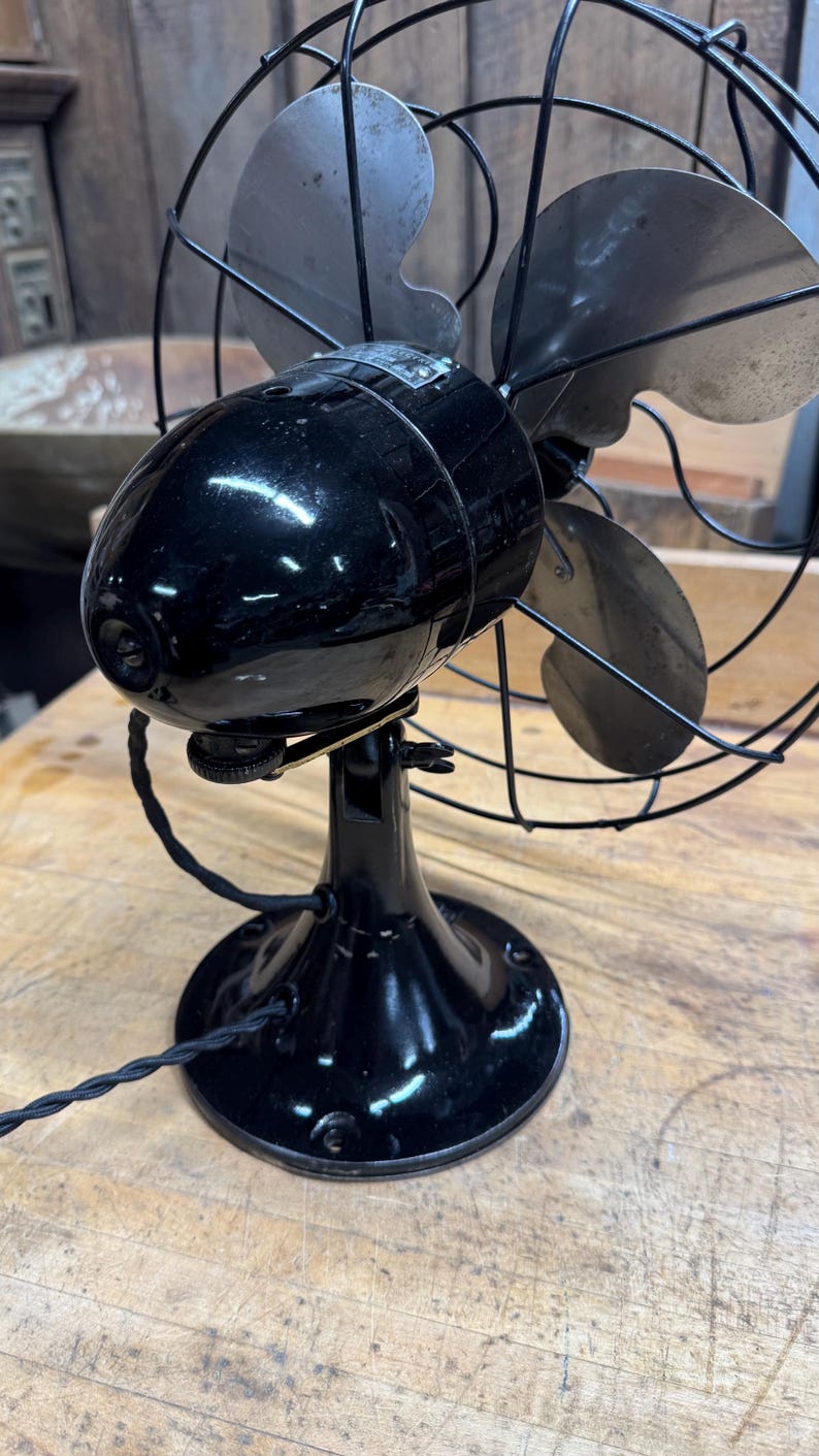 May include: Vintage black electric fan with three silver blades, enclosed by a protective wire cage. The fan has a glossy finish and a round base. A black power cord extends from the base. The fan is a classic home appliance.