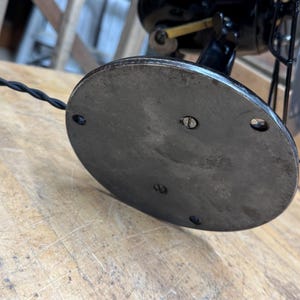 May include: Close-up of the base of a vintage black electric fan. The base is a dark, oval-shaped metal plate with screw holes. A black, braided power cord with a green tie is visible. The fan's cage and blades are also visible.