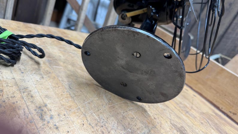 May include: Close-up of the base of a vintage black electric fan. The base is a dark, oval-shaped metal plate with screw holes. A black, braided power cord with a green tie is visible. The fan's cage and blades are also visible.