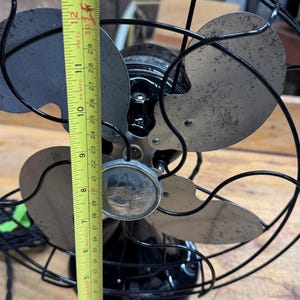 May include: Vintage Emerson Electric fan, black with silver blades, approximately 12 inches tall. The fan has a protective wire cage and a decorative emblem on the front. The blades show signs of wear.