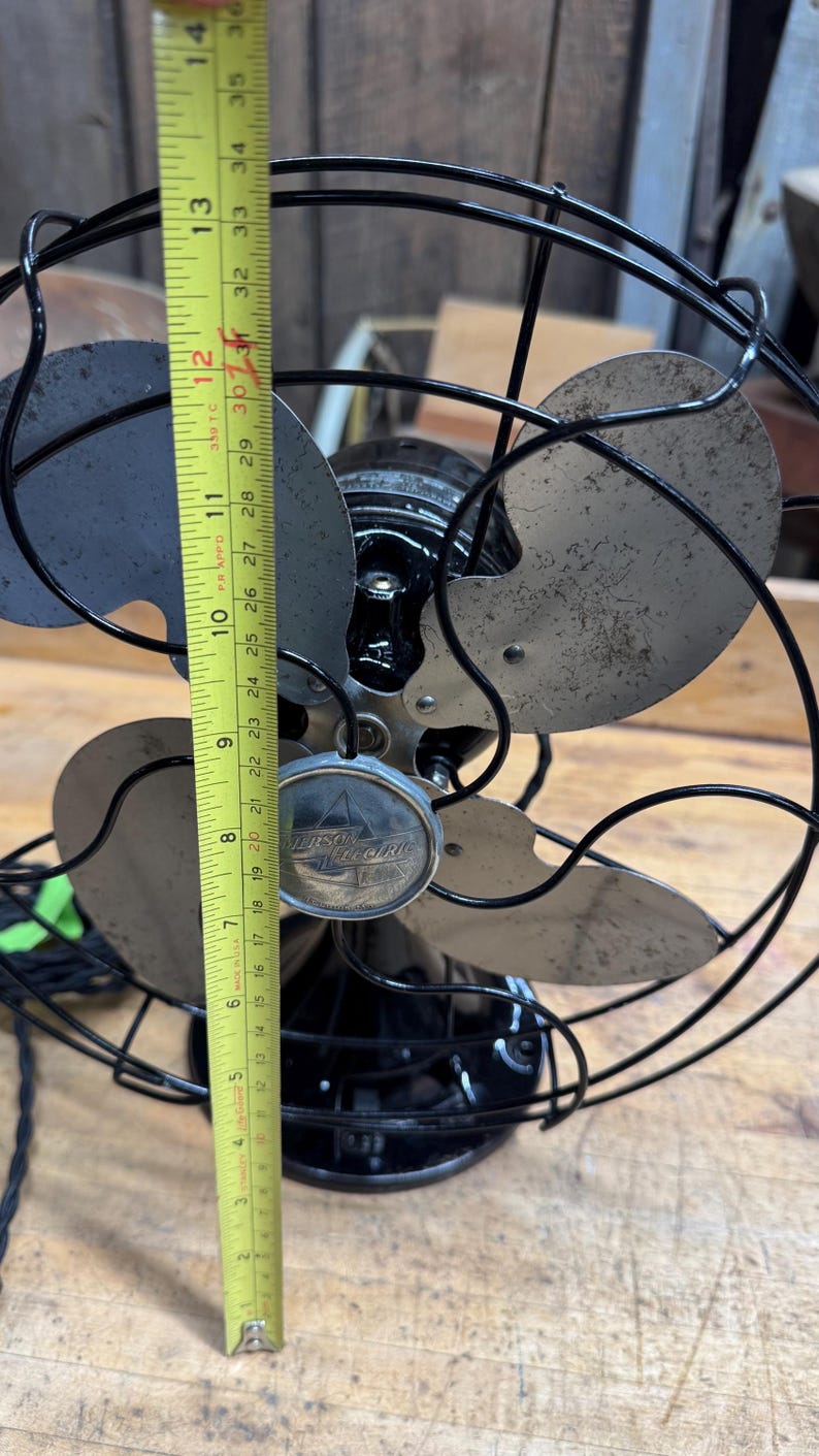May include: Vintage Emerson Electric fan, black with silver blades, approximately 12 inches tall. The fan has a protective wire cage and a decorative emblem on the front. The blades show signs of wear.
