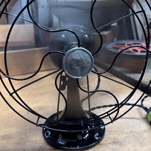 May include: An antique Emerson Electric fan, featuring a black metal cage and base. The fan's blades are visible behind the cage, and the Emerson Electric logo is displayed on the front. The fan is positioned on a wooden surface.