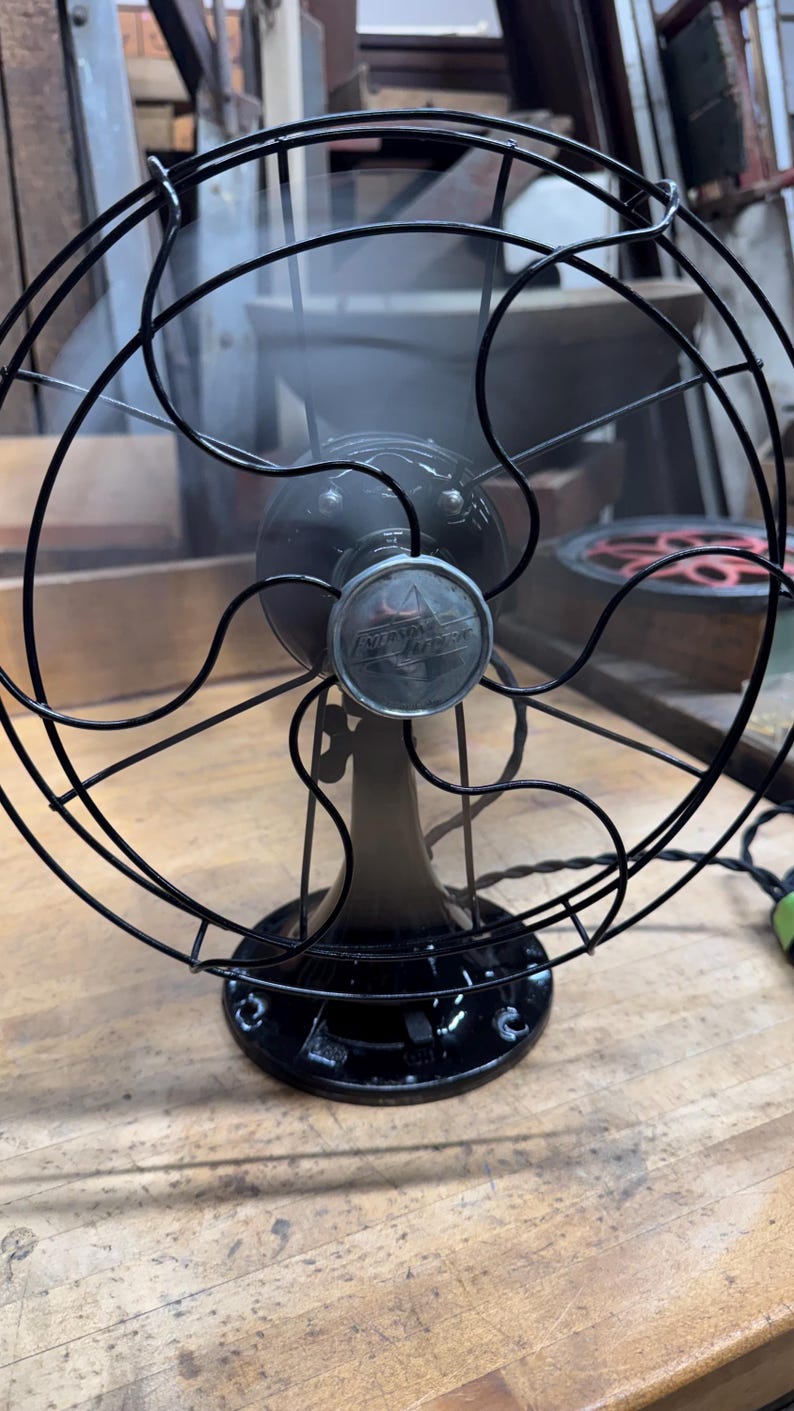 May include: An antique Emerson Electric fan, featuring a black metal cage and base. The fan's blades are visible behind the cage, and the Emerson Electric logo is displayed on the front. The fan is positioned on a wooden surface.